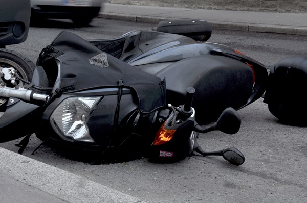 Black motorcycle lying on its side on a city street near the curb, showing signs of crash damage with its headlight exposed and turn signal illuminated.