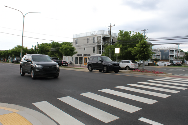 An urban intersection with a wide crosswalk shows multiple updated vehicles, including a dark gray SUV positioned near the crosswalk. The scene is clear of people, with trees, buildings, and power lines in the background under a cloudy sky.