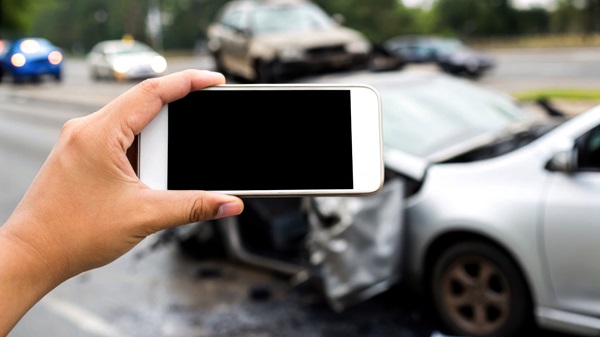 A hand holds a smartphone horizontally, taking a picture of a silver car with front-end damage and a black screen visible on the phone, with other damaged vehicles blurred in the background on the road.