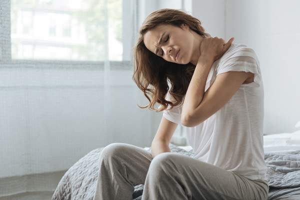 A woman sitting on the edge of a bed, wearing a white t-shirt and grey lounge pants, winces in pain while clutching her neck and shoulder.