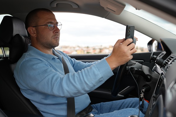 Driver holding a smartphone with both hands while seated behind the steering wheel of a passenger vehicle, illustrating distracted driving subject to fines and penalties