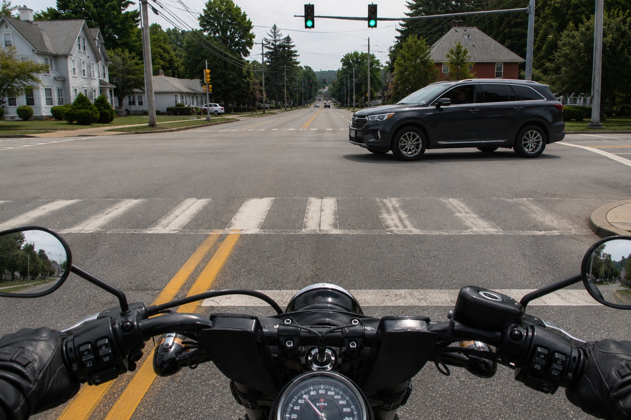 A first-person view from a motorcycle stopped at a suburban intersection, showing an SUV turning left across the rider's path, which illustrates the common right-of-way hazards that contribute to motorcycle accident risks in New Hampshire.