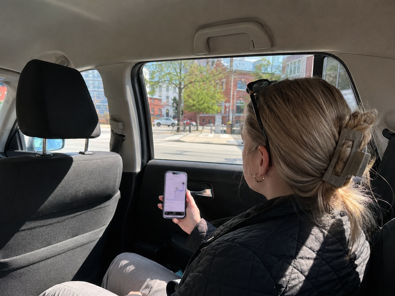A female rideshare passenger sits in the back seat of an Uber or Lyft vehicle in New Hampshire, viewing a navigation route on her phone while passing through a downtown area, depicting the insurance coverage questions that arise for passengers injured during an active rideshare trip under New Hampshire's rideshare insurance framework.